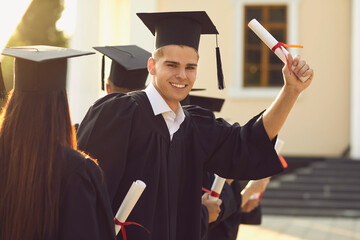 Young smiling boy university graduate in black bonet and mantle standing and holding dimpoma with...
