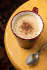 Aerial closeup of cappuccino coffee cup and spoon on wooden stool, on black background, in vertical