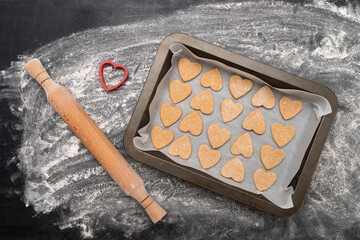 Wooden rolling pin and baking sheet with heart-shaped cookies. Homemade baking on black table