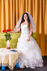A Jewish bride with black curls in a white dress with a veil stands in the room waiting for the groom for the chuppah ceremony.