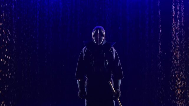 Japanese kendo fighter lowering his shinai towards camera. Athlete in kimono, martial and with bamboo sword poses against dark studio background with blue light and rain. Close up. Slow motion.