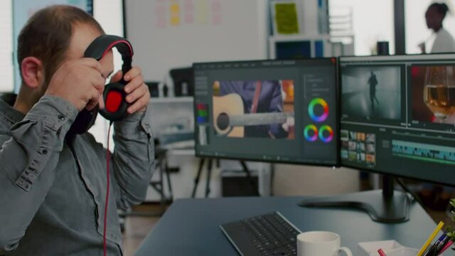 Videographer editing video project putting on headphones working in start up creative agency office. Man processing film montage in digital multimedia company using post production software