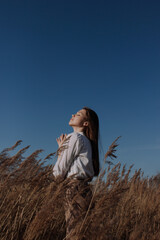 Young woman with closed eyes in white blouse standing in field of dry pampas grass in front of blue sky and sun. Urban style and street fashion. Girl in casual outfit looking up in the sky
