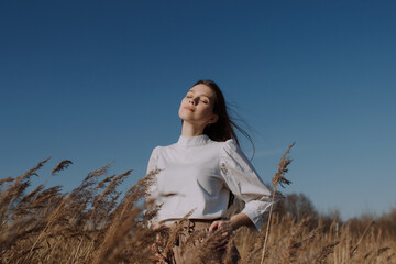 Young woman with closed eyes in white blouse standing in field of dry pampas grass in front of blue...