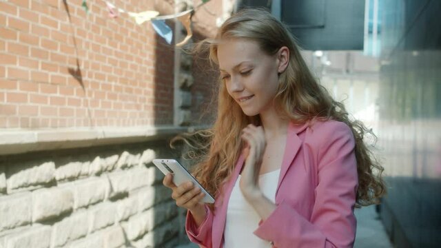 Slow motion of beautiful lady using smartphone touching screen laughing standing in city street alone on windy day. Positive emotions and devices concept.