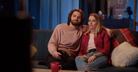 Obraz premium Young beautiful woman smiling in living room when her boyfriend arrives home. Joyful affecitonate couple lovers watching Tv on evening. Nighttime.