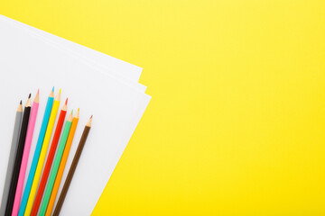 Many new colorful pencils and white paper on bright yellow table background. Closeup. Empty place for text. Top down view.