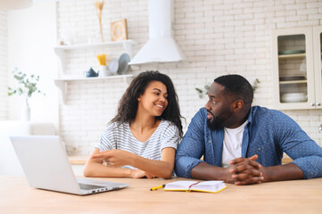 Excited young black couple or friends watching online video classes blog, sitting in the kitchen,...