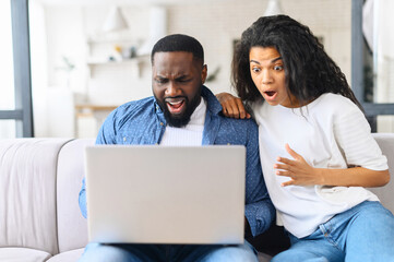 Vexed and surprised African-American couple sits on the couch at home and looks at the laptop screen, received unexpected news, lost in video game