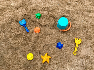 Fototapeta na wymiar Top view of bright colored plastic molds, shovel, rake and bucket in the sandbox, against the background of dark sand with raindrops. Copy space