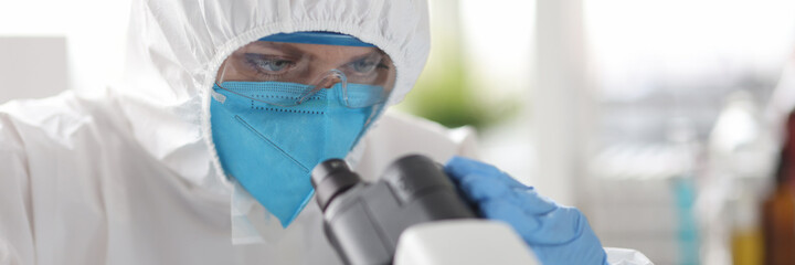 Scientist in protective suit and respirator looking through microscope in laboratory