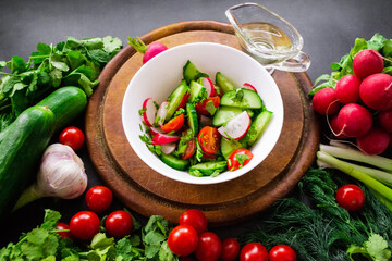 A pile of fresh, ripe vegetables: radishes, tomatoes, cucumbers, herbs, and garlic are arranged around a round wooden chopping board. A white bowl with vegetables cut into a salad and olive oil