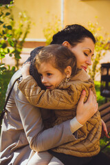 A little girl with dark hair in a mustard-colored jacket hugs her mother sitting on bench in a park. Mom embracing her lovely daughter on the warm autumn day. Motherhood. Happy mothers day