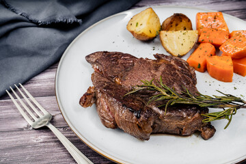 Beef steak served on a plate with rustic potatoes and carrots on a wooden table. 45 degree view