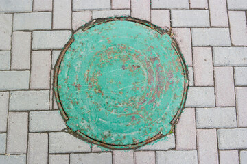 Old iron manhole cover embedded with flaked green paint, mounted in the paving stones, top view