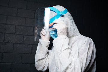 Close-up portrait of young doctor man wearing PPE suit against coronavirus and covid-19. Background of black brick wall. Putting on safety goggles.