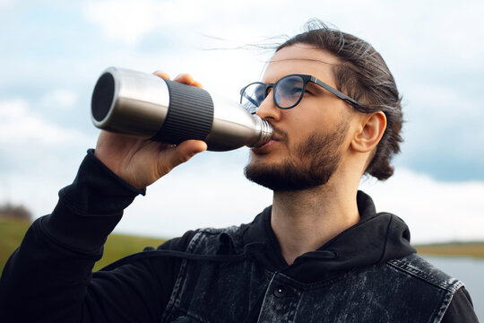 Portrait Of Young Man Drinking Water From Reusable Metal Bottle On Background Of Blurred Cloudy Sky.