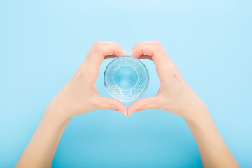 Heart shape created from young adult woman hands around glass of fresh water on light blue table background. Pastel color. Closeup. Point of view shot. Healthy drink concept. Top down view.