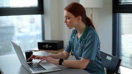 Attractive young female practitioner doctor wearing blue green medical uniform working typing on laptop sitting at desk near window in modern office of medic clinic. Shooting in slow motion.