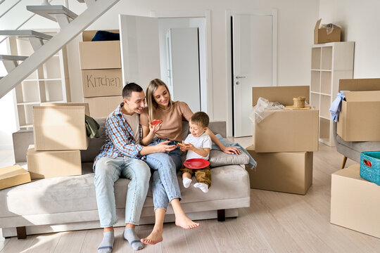 Happy young parents sitting on sofa with kid relaxing in new home in relocation. Joyful family in living room at moving day, boxes on background, mom and dad happy new householders tenants concept. - Powered by Adobe