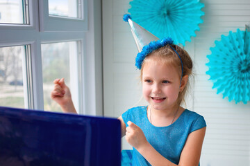 A little girl, curves and laughs in front of a laptop in a cap. Celebrates birthday via internet in...