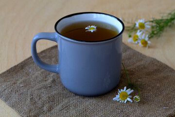 hot drinks. grey mug of chamomile tea on sackcloth. cup of hot Herbal chamomile tea on a wooden table with copy space with fresh flowers and green leaves on light background.