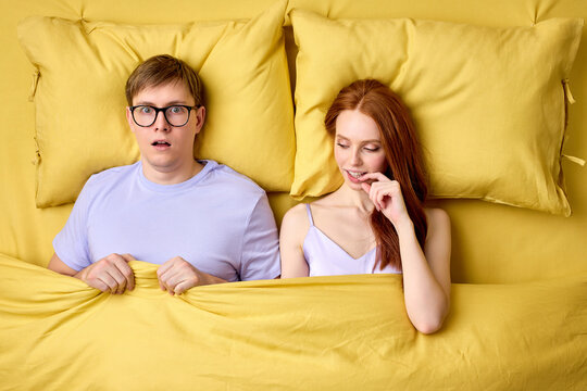Happy couple lying in bed under duvet after first sex experience, man is surprised by skills of female in bed