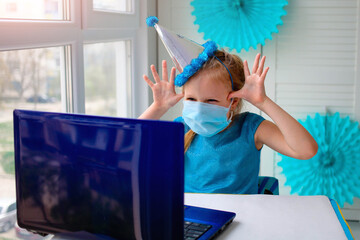 A happy little girl, crooked in a medical mask in front of a laptop celebrates a birthday via the...