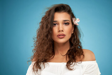 Fototapeta premium Front view portrait of attractive brunette woman with curly hair and in white sweater on blue background. Concept of modern shooting on professional photo studio. 
