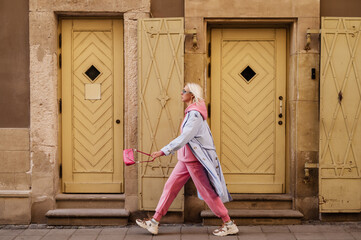 Street style, fashion: woman wearing  trendy sport chic outfit walking in street of European city. Blue trench coat, sunglasses, pink hoodie, trousers, sneakers, mini bag. Copy, empty space for text - 429289039