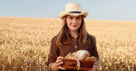 Portrait of happy Caucasian female farmer standing in golden field with basket of bread and baking in hands. Joyful girl in hat smiling to camera in countryside on sky background. Farming concept