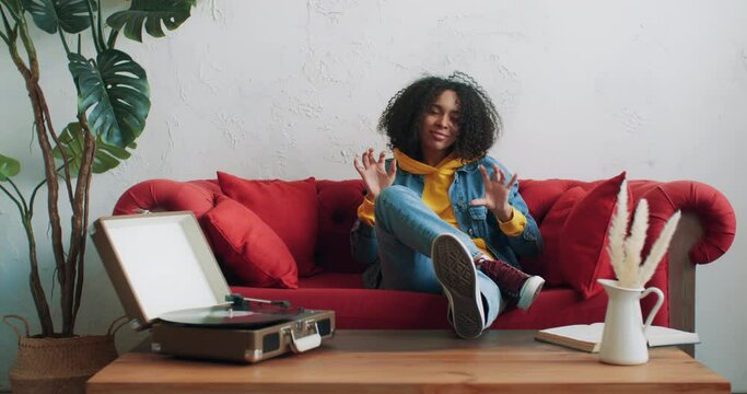 Happy stylish african american black female in yellow hoodie and jeans jacket listening music on vinyl turntable vintage record player dancing sitting on red sofa at home. Gen z cheerful urban woman