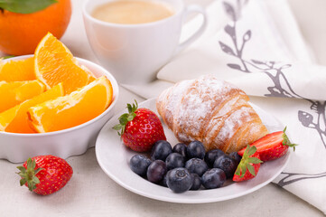 Breakfast with a cup of coffee, fresh croissant and ripe strawberries and blueberries and juicy oranges on a white background