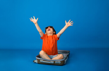 happy little boy in an orange T-shirt and denim sits in a suitcase on a blue background with space...