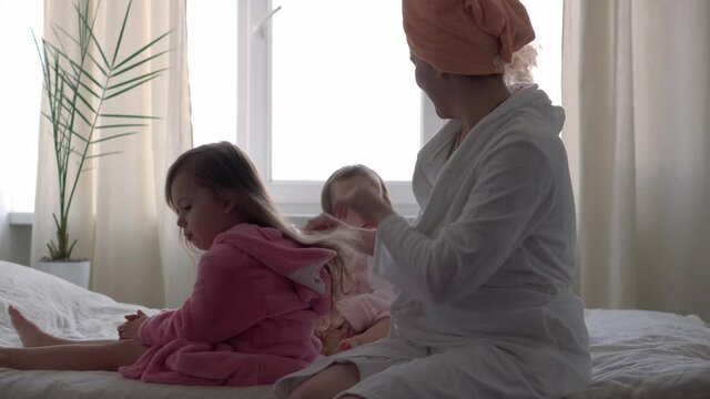 Authentic cute young woman combing long hair of baby girl. mom with 2 children daughters different ages in bathrobes after bath on white bed oposite window. Spa, motherhood, childhood, family concept.