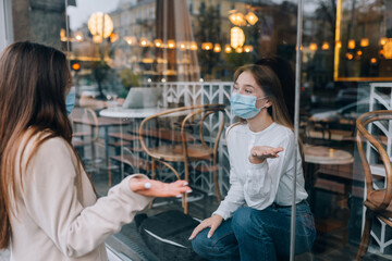 Two women in protective masks opposite each other, window between them.