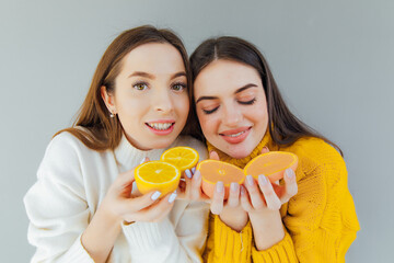 Two pretty charming joyful attractive cheerful funny comic positive nude natural pure girls having two pieces of orange, closing one eye, isolated on gray background