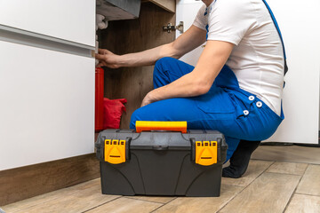 Handyman in overalls sits near the sink in the kitchen and looks at what is the cause of the broken pipes, there is a box of tools next to it