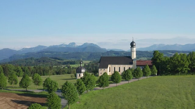 Church of pilgrimage in Wilparting Irschenberg during summer, mountains in background, blue sky, Bavaria Germany, drone view flying backwards
