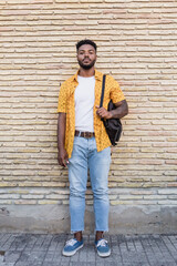 Lifestyle portrait of an attractive american young man with beard smiling and carrying a backpack with a brick wall as background