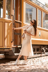 Full length portrait. Young woman in beige blouse, skirt and white sneakers walking at town street...