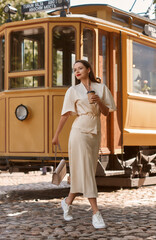Young beautiful woman in beige skirt and blouse walking old town street with pavement on a summer sunny day. Pretty girl with cup of coffee standing against retro tram