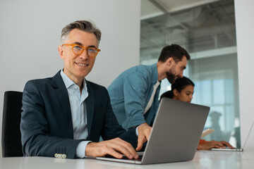 Confident mature business man wearing stylish eyeglasses using laptop computer looking at camera. Group of business people working together in modern office 