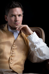 A handsome Regency gentleman sitting in a red velvet chair in a darkened room