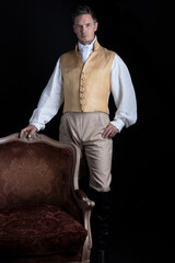 A handsome Regency gentleman standing beside a red velvet chair in a darkened room 