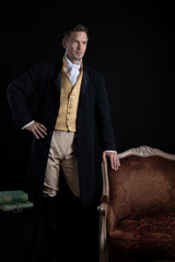A handsome Regency gentleman standing beside a red velvet chair in a darkened room 