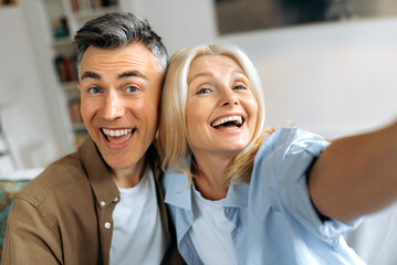 Close-up headshot of happy cute funny mature spouses in casual clothes, Caucasian husband and wife taking selfie on smartphone in their living room, looking at camera, smiling, spend leisure time