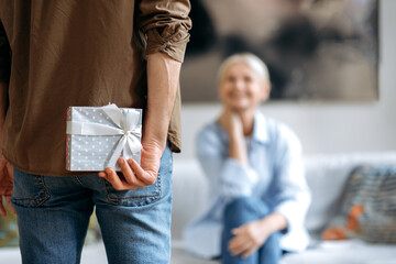 A present from husband on a special day. A loving husband in casual stylish clothes, holds a beautiful small box behind his back, prepares to give a surprise his beloved wife, a small anniversary gift