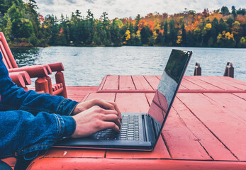 Young professional - digital nomad taking advantage of outdoors in autumn - working on laptop by the edge of a lake. Blurred background - autumn colors