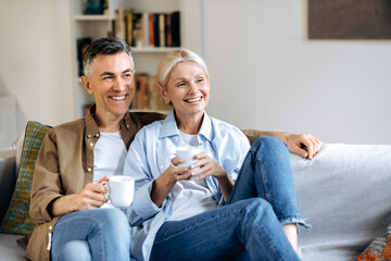 Glad mature caucasian married couple in stylish casual clothes, sitting on a comfortable sofa in the living room, holding cups of tea, chatting, spending time at home, looking away, smiling